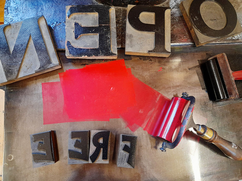 A paint roller with a swipe of red paint, surrounded by print blocks spelling 'OPEN' and 'FREE' on a table