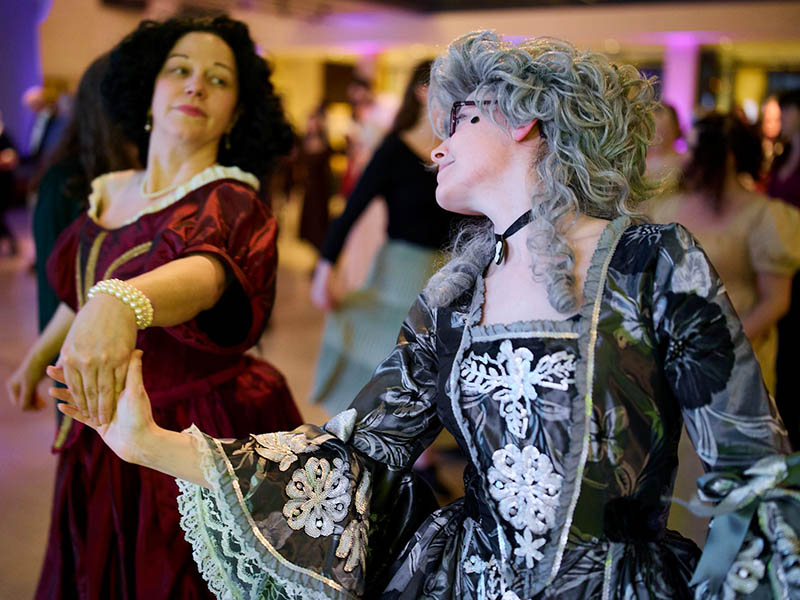 Two people wearing vividly coloured Regency dress are dancing in a hall