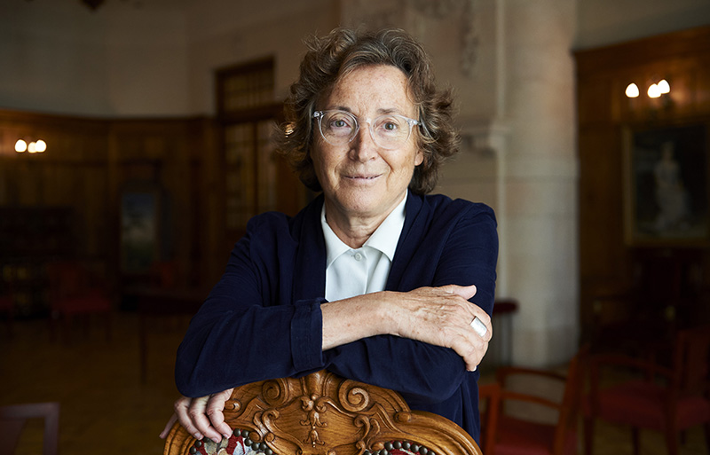 A woman with short hair wearing glasses, a white blouse and dark jacket leans against the back of an ornate wooden chair