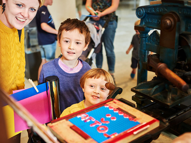 A woman wearing a yellow jumper and two children stand in front of a historical printing press, which has been set up with LEGO