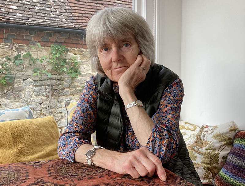 A woman with grey hair sits at a table; she is wearing a floral shirt and a dark gilet
