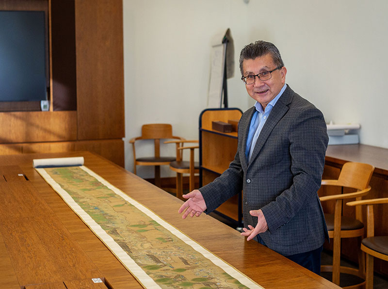 A man wearing a dark suit and blue shirt stands next to a long table on which is a long painted scroll