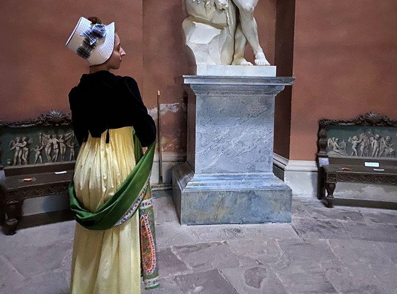A woman wearing a yellow dress with a dark jacket, green sash and white hat stands in a gallery in front of a marble statue