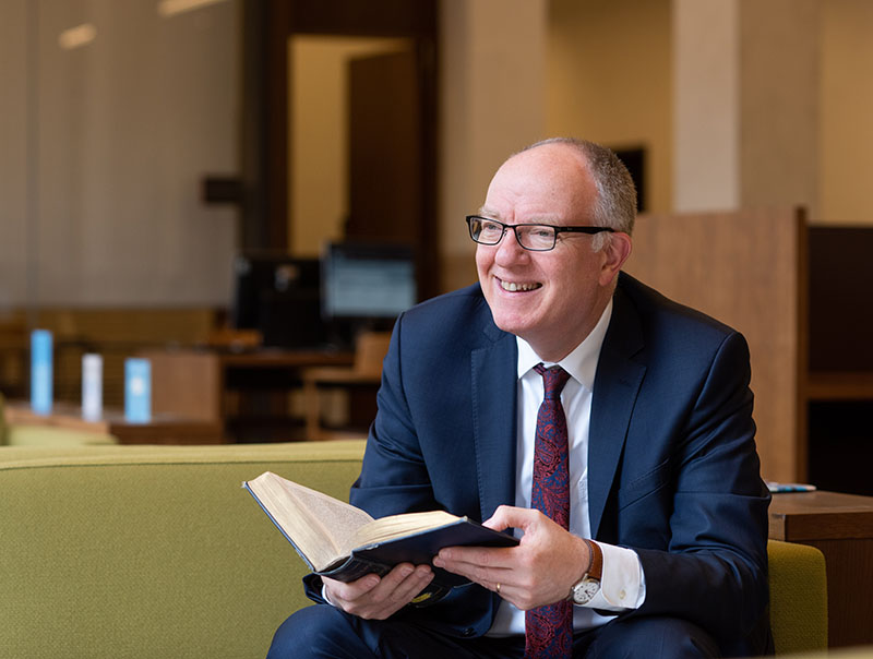 A man wearing a dark blue suit sits on a green sofa, holding an open book