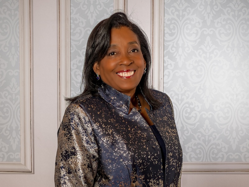 A Black woman with dark hair wearing a purple and silver jacket stands against a panelled wall with blue wallpaper