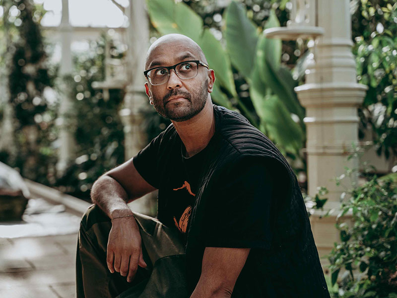 A man wearing dark clothes and glasses kneels in a garden area with plants around him