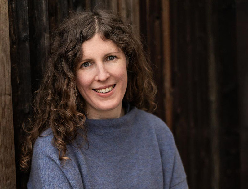 A woman with dark curly hair wearing a blue jumper stands against a dark wooden background