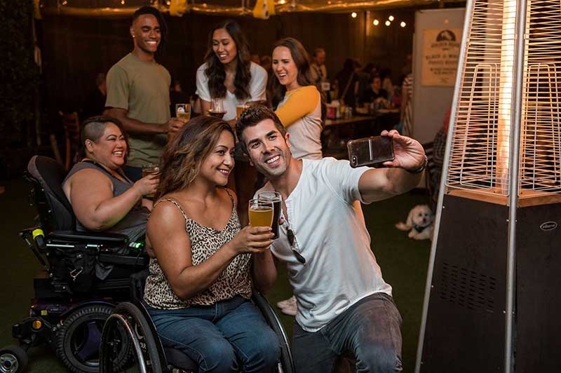 A man and a woman in a wheelchair are taking a selfie together while the other friends—three standing, one in a wheelchair—smile in the background.