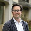 A man with dark hair and glasses wearing a jacket and shirt stands outside
