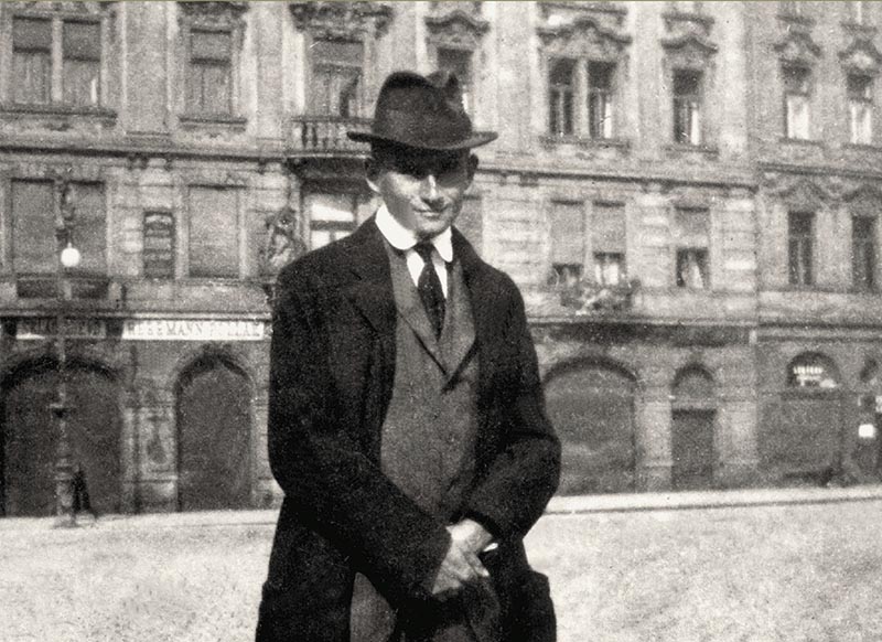 A black-and-white photograph of Franz Kafka standing in front of a building; he wears a dark suit and hat