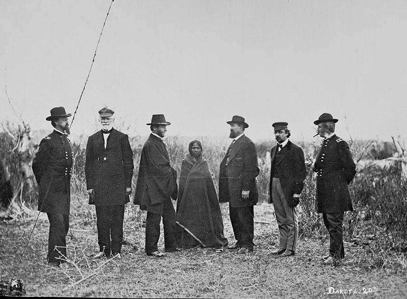 A black and white photograph of six men in 19th-century period dress standing side-by-side with a girl wrapped in a blanket