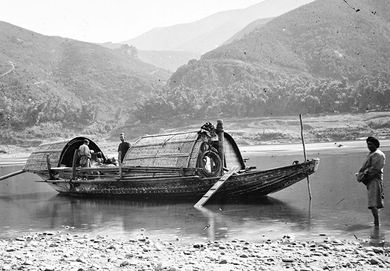 A black and white photograph of a traditional flat-bottomed wooden boat on a river