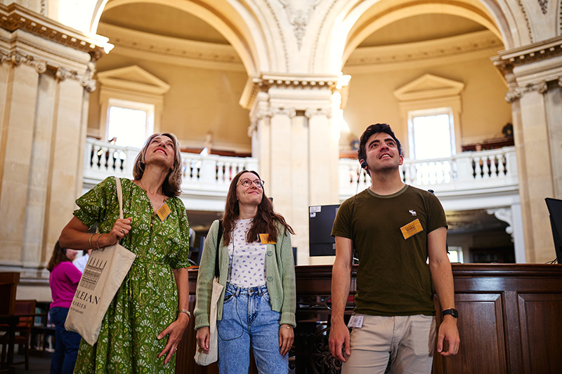 Three people stand in the middle of the Radcliffe Camera, looking up; they are in front of large stone arches