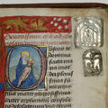 A detail of an illuminated letter showing a woman in blue holding the body of a male person. On the right of the page are two silver badges pinned to the page