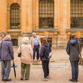 A tour group wearing coats stands in front of a building made of warm-coloured stone, listening to a tour guide