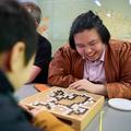 A man wearing a brown jacket smiles as he looks at a go board with white and black stones arranged on it