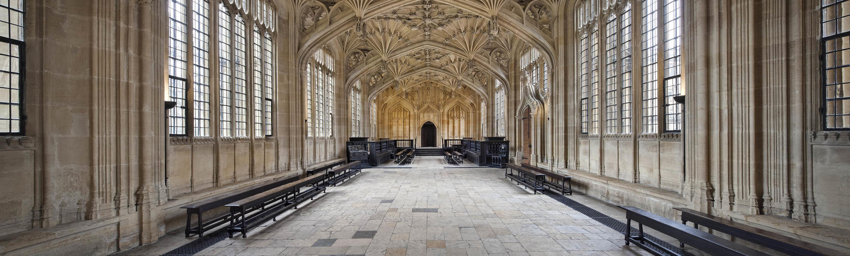 View of the Divinity School in the Bodleian Library, a 15th-century room with ornate stone ceiling carvings, a stone floor, and windows down either side of the room