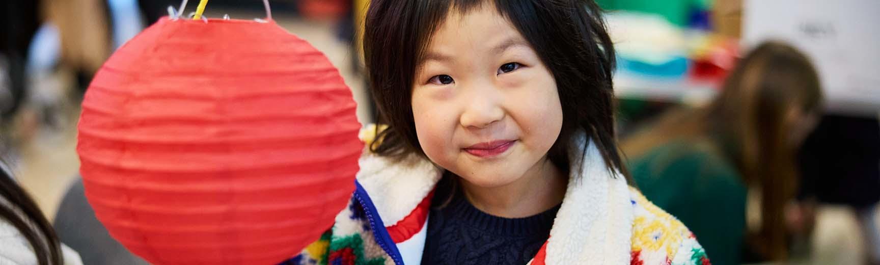A girl with dark hair wearing a brightly coloured fleece holds up a red lantern 