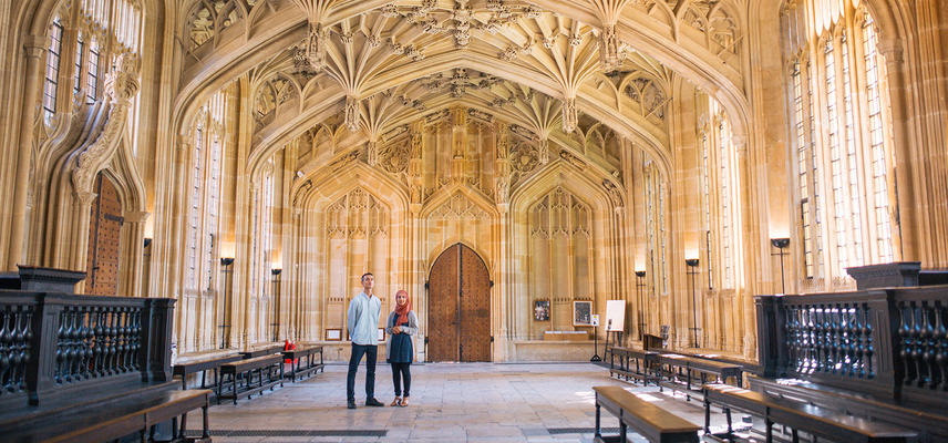Two people stand in a large room with an ornately carved stone ceiling