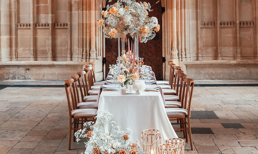 A flower setting with pale pink and pale orange flowers on a table set with candles, wine glasses and china plates