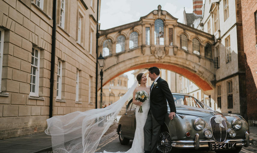 A couple in their bridal outfits stand in front of a vintage silver car underneath the Bridge of Sighs in Oxford