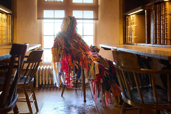A person dressed in a whole body costume of material strips sits in a chair in an old library
