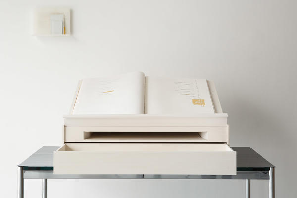 A white book open on a white lecture on a table, the two pages have poetry on either side and on the wall behind is a white shelf with another white object on it