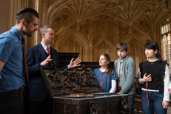 A tour guide stands behind a wooden chest, a group stands around him listening