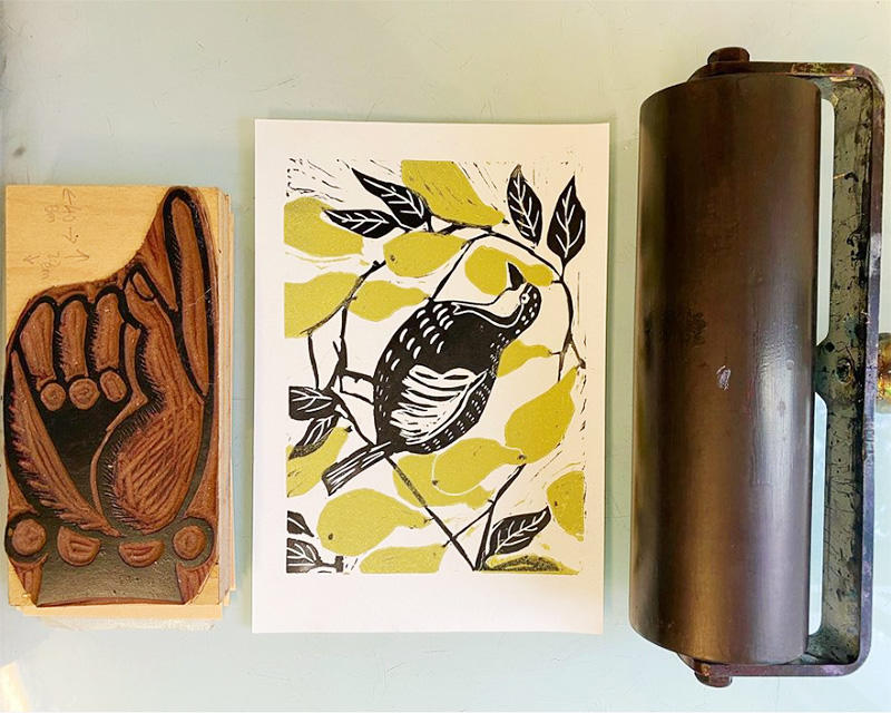 A printing block of a hand, a linocut print of a bird amongst yellow leaves, and printing equipment