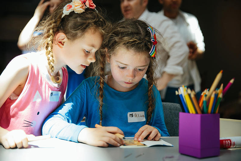 A girl in a blue jumper colours in a drawing while another girl in pink looks over her shoulder
