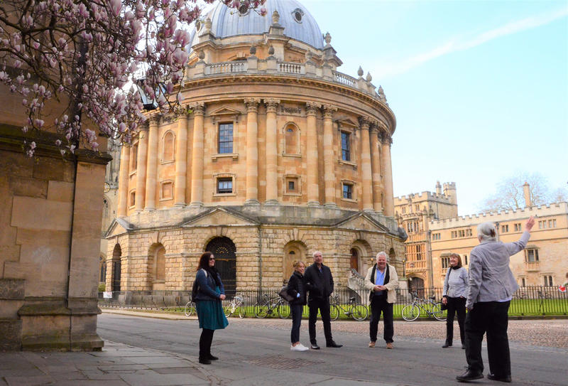 A group of five people look towards a tour guide who is point out a historic building detail, in the background is the Radcliffe Camera