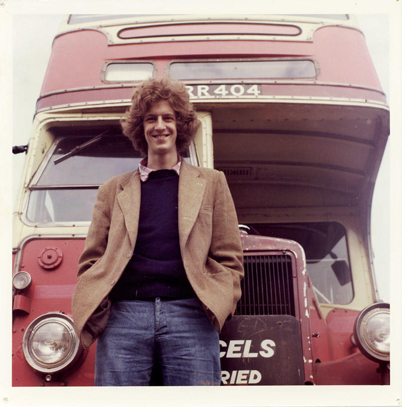 A photograph of a man wearing a jacket and jeans standing in front of a red double-decker bus, the Free Photographic Omnibus